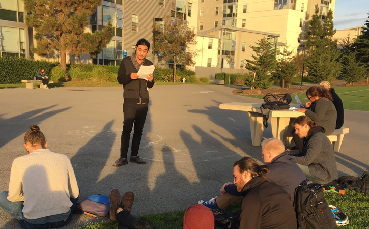 A graduate student presenting their thesis to a group of other students outside.