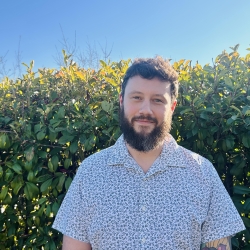 Photo of Jacob Holland standing in front of a hedge in the sun.