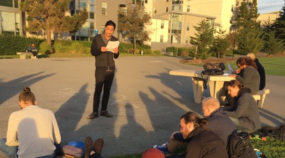 A graduate student presenting their thesis to a group of other students outside.